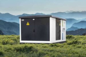 Eine Trafostation mit weißen Wänden und anthrazitfarbenen Türen steht auf einer grünen Wiese, umgeben von einer beeindruckenden Berglandschaft im Hintergrund. Die moderne Kompaktstation ist ideal für Photovoltaik, E-Mobilität, Windkraft und Netzbetreiber.